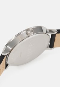 Montre en acier inoxydable argenté avec couronne texturée, bracelet noir et nom de la marque gravé au dos lisse.