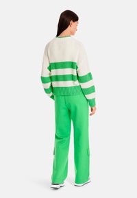 Weißer Strickpullover mit grünen Streifen, kombiniert mit grünen weit geschnittenen Hosen. Der Pullover hat einen gerippten Saum und einen lässigen Schnitt sowie einen strukturierten Stoff.