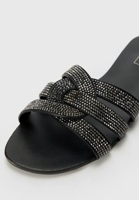 Crne natikače s preklopljenim trakama ukrašenim srebrnim rhinestones, teksturiranim gornjim dijelom i glatkim, ravnim đonom.