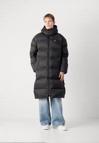 Sort pufferjakke med høj krave, fuld lynlås og vandret quiltet design. Båret over lyseblå flared jeans og hvide sneakers.