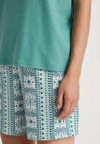 Korte mouwen teal top, gecombineerd met een patroonshort in turquoise en wit, met geometrische vormen en palmmotieven.