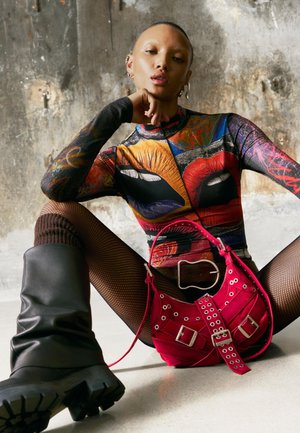 Persoană purtând un top colorat cu model de buze, plasă pentru picioare, cizme negre și o centură mare neagră, ținând o geantă roșie cu catarame.