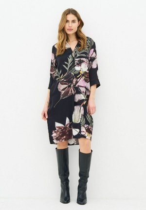 Woman wearing a knee-length black dress with large pink and white floral prints and black knee-high boots, standing against a white background.