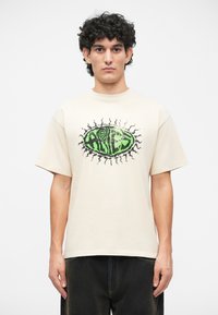 Tricou bej cu mânecă scurtă, având un design grafic mare de culoare verde cu accente negre, în formă de ochi. Croială relaxată și stil cu gât rotund.