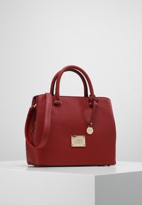 Röd läderhandväska med en strukturerad yta, dubbla handtag i toppen och en justerbar axelrem. Har en metallisk varumärkesplatta.