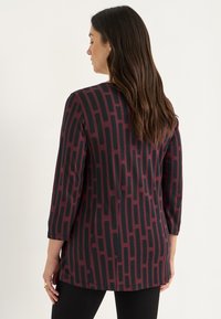 Vrouw met lang bruin haar, gekleed in een maroon en zwart gepatterde lange mouwtop, die zijwaarts tegen een effen lichte achtergrond staat.