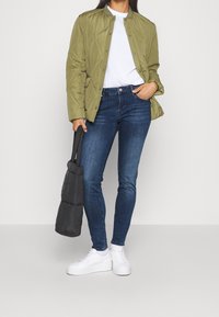 Vihreä oliivinvärinen tikattu takki, jossa on napit, valkoinen t-paita, tummansiniset skinny-farkut ja valkoiset lenkkarit. Musta puffikassi kädessä.