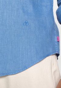 Camisa de lino azul claro con dobladillo curvado, cuenta con un logo bordado y una etiqueta de marca rosa en el costado. Textura suave, diseño informal.