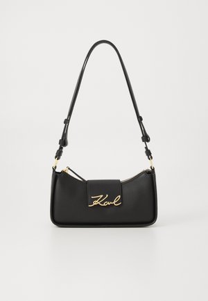 Svart skinnveske med buet design, som har et gullfarget signaturlogo foran, glidelås-lukking og justerbar stropp.