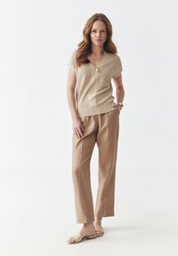 Beige pletený top s krátkým rukávem a výstřihem do V, kombinovaný se světle hnědými lněnými kalhotami. Modelka má na sobě neutrální sandály a jednoduchý zlatý náhrdelník.