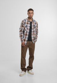 Camisa de cuadros en marrón, crema y negro; con dos bolsillos frontales; combinada con pantalones marrones oscuros y zapatillas blancas con detalles en crema.