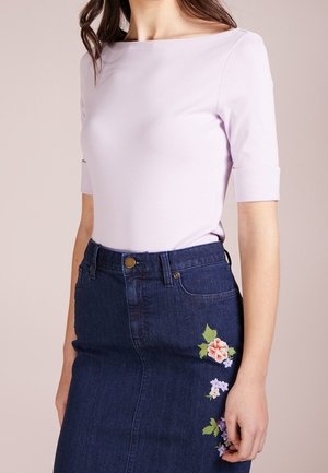 Femme portant un haut violet clair à manches coudées rentré dans une jupe en denim foncé avec une broderie florale sur le côté droit.