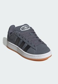 Zapatilla de ante gris con tres rayas negras, logo redondo en la lengüeta, cordones grises, entresuela blanca y suela de goma marrón.