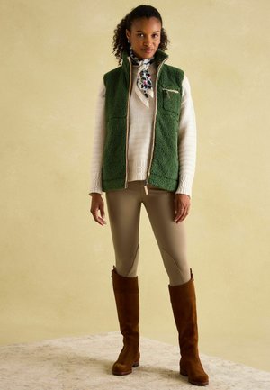 Grüne Fleece-Weste mit Reißverschlusstasche, cremiger strukturierter Pullover, beige Leggings und braune kniehohe Stiefel. Weicher Stoff und schichtige Gestaltung.