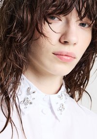 Camicia bianca con colletto gioiello impreziosito da strass trasparenti, tessuto morbido e design con bottoni. I capelli appaiono leggermente bagnati e disordinati.
