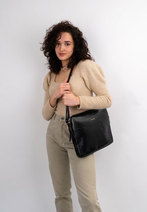 Black crocodile-embossed handbag with a gold zipper and shoulder strap. Smooth texture and rectangular shape, held by a person.