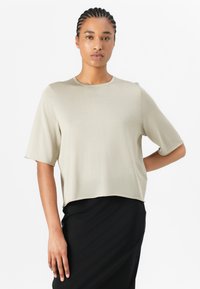 Short-sleeve, beige top made from a smooth fabric with a round neckline. The design features a loose fit and subtle stitching details.