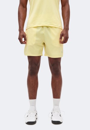 Homme portant un short de sport jaune pâle et un t-shirt assorti, des chaussettes blanches et des baskets blanches Adidas, debout devant un fond uni.