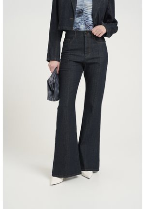 Frau trägt dunkle ausgestellte Jeans, kurze Jeansjacke, gerafftes blaues Oberteil, hält eine graue Clutch und weiße spitze High Heels.