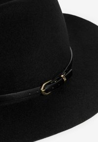 Čierny filcový klobúk so širokým okrajom, s tenkým čiernym koženým pásikom a malou zlatou prackou na bielom pozadí.