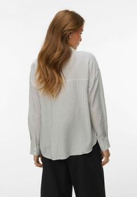 Femme aux cheveux longs et ondulés, portant une chemise ample à manches longues rayée verticalement blanche et noire, ainsi qu'un pantalon noir à jambes larges, vue de dos.