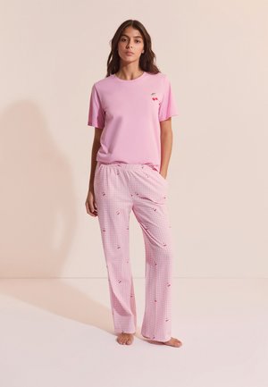 Femme debout pieds nus, portant un t-shirt rose à manches courtes avec un petit motif de cerise et un pantalon à carreaux roses avec des imprimés de cerises.