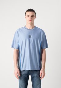 Light blue short-sleeve sports shirt with a round neckline, textured fabric, and contrasting blue side piping. Features a logo on the chest.