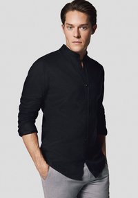 Czarna koszula na guziki z stójką, długimi rękawami i luźnym krojem. Tkanina wydaje się gładka z matowym wykończeniem. Model stoi z rękami w kieszeniach.