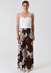 Top bianco senza maniche con scollatura a cuore, abbinato a pantaloni larghi a fantasia floreale marroni in un tessuto morbido. Accenti di gioielli semplici.