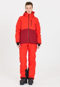 Rote und kastanienbraune Skijacke mit Frontreißverschluss, Kapuze und seitlichen Taschen, kombiniert mit passenden roten Hosen und schwarzen Skischuhen.