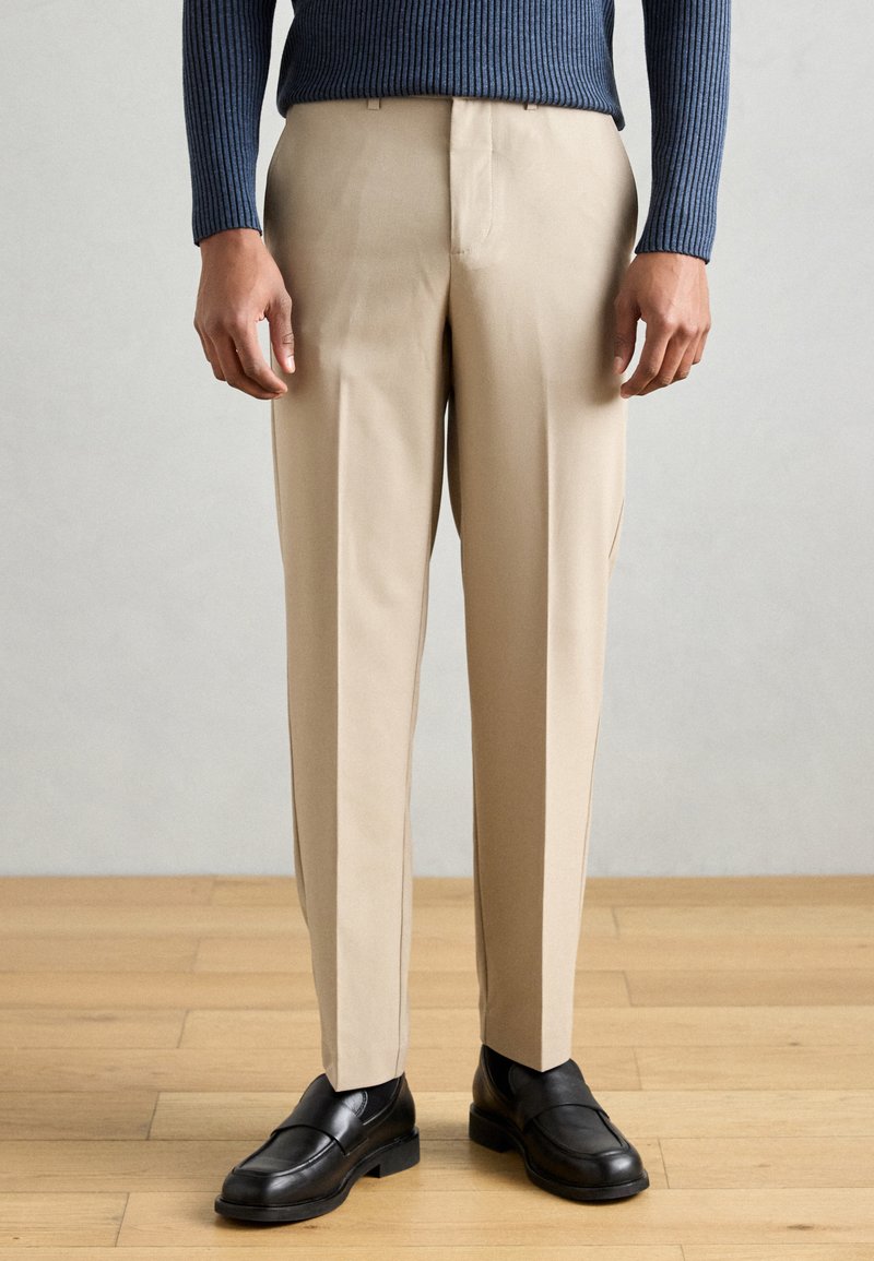 Bézs, testre szabott szabású nadrág, sima textúrával és oldalzsebekkel, fekete mokaszinokkal egy fa padlós háttér előtt.