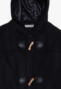 Giacca nera con cappuccio, chiusure a fibbia in legno e dettagli in gomma. Tessuto morbido, fodera in satin liscia, design classico adatto ai bambini.