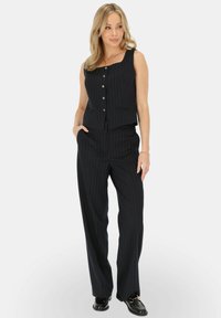 Czarny garnitur w paski składający się z bezrękawnika z kwadratowym dekoltem i zapięciem na guziki, połączony z szerokimi spodniami i czarnymi butami.