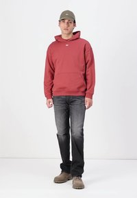 Red hoodie with a kangaroo pocket and an embroidered logo, paired with dark jeans and beige shoes. Man stands against a white background.