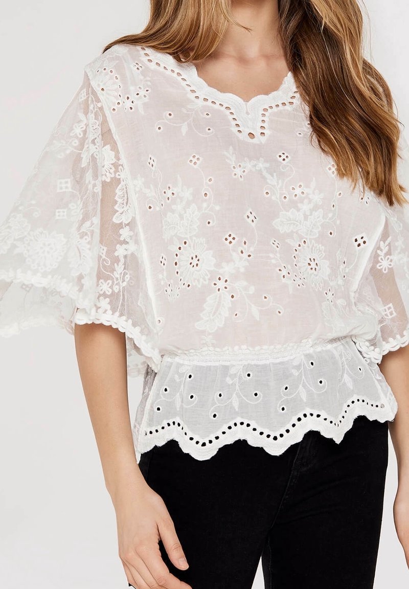 White embroidered blouse with lace accents, floral patterns, and scalloped hem; featuring loose, flowing sleeves and a v-neck design.