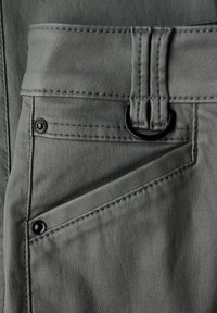 Close-up van een zak van een grijze broek met zwarte klinknagels en een stoffen riemlus waarin een zwarte D-ring zit.