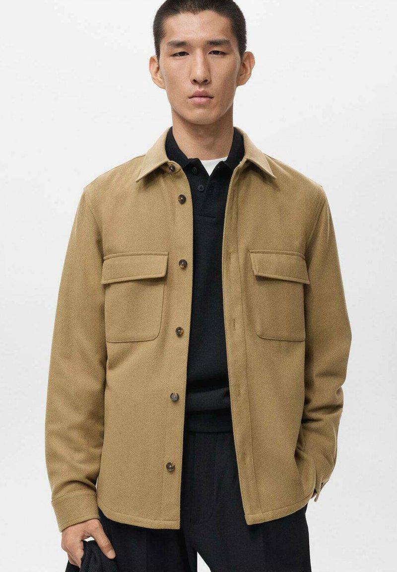 Tan wool jacket with button closures, two front pockets, and a classic collar, layered over a black polo shirt. Smooth texture, tailored fit.