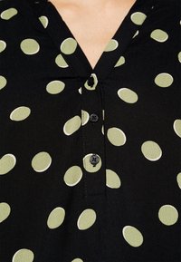 Mustat pusero, joka on valmistettu kankaasta ja jossa on V-pääntie. Puserossa on suuria vaaleanvihreitä ja valkoisia pilkkuja, sekä mustia nappeja edessä.