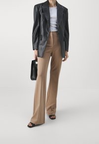 Blazer noir en similicuir à texture crocodile, cintré aux épaules. Pantalon beige taille haute, jambes larges, associé à des sandales et un sac noirs.