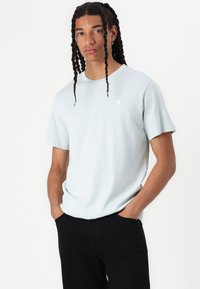 Hellblaues Baumwoll-T-Shirt mit kurzen Ärmeln, Rundhalsausschnitt und dezentem weißen Logo auf der Brust. Kombiniert mit schwarzen Hosen.