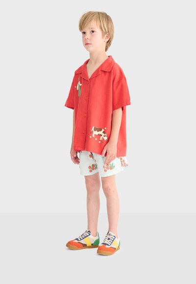 Enfant blond debout, portant une chemise rouge à manches courtes avec des motifs de vaches et de plantes, un short blanc avec des motifs de fruits orange, et des baskets colorées.