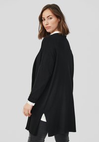 Czarny kardigan z długim rękawem o luźnym kroju, wykonany z miękkiego materiału, z bocznymi rozporkami i zaokrąglonym dołem, noszony na białej koszuli.