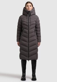 Frau, die auf einem hellen Hintergrund steht, trägt einen langen grauen gefütterten Wintermantel mit hohem Kragen und schwarzen, kniehohen Stiefeln.