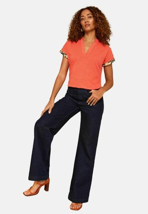 Nainen kiharat hiukset, päällään korallinvärinen lyhythihainen toppi leopardikuvioisilla kalvosimilla, tummat leveälahkeiset housut ja oranssit korkokengät, poseeraa itsevarmasti.