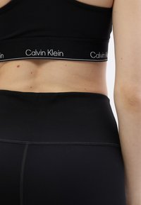 Melna sporta krūšturis ar kontrastējošu baltu Calvin Klein logotipa joslu, apvienots ar augstās jostasvietas melnām legingām ar tekstūru šuvēm.
