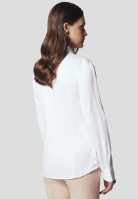 Biała koszula z długim rękawem, z kołnierzykiem zapinanym na guziki, o gładkiej fakturze, dopasowanym kroju i zaokrąglonym dolnym brzegu, noszona z beżowymi spodniami.