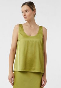 Vrouw in een mouwloze satijnen olijfgroene top met een ronde hals, gecombineerd met een bijpassende olijfgroene rok, tegen een effen achtergrond.