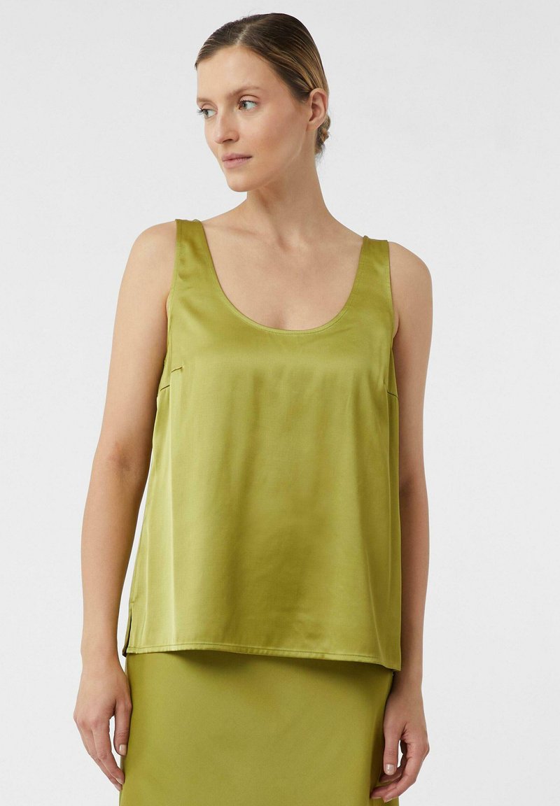 Vrouw in een mouwloze satijnen olijfgroene top met een ronde hals, gecombineerd met een bijpassende olijfgroene rok, tegen een effen achtergrond.