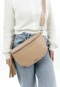 Riñonera de cuero beige con forma curva, cremallera frontal y detalle de borlas, llevada sobre un suéter blanco de punto trenzado y jeans claros.
