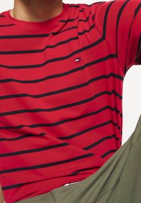 Rød kortærmet T-shirt med sorte vandrette striber og et lille logo nær brystet. Lavet af blødt stof og med rund hals.